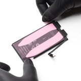 A replacement smartphone battery with a pink protective film and black connector tabs, held by hands wearing black gloves on a white background.