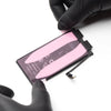 A replacement smartphone battery with a pink protective film and black connector tabs, held by hands wearing black gloves on a white background.