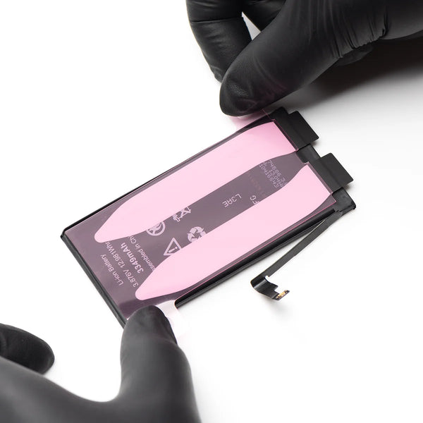 A replacement smartphone battery with a pink protective film and black connector tabs, held by hands wearing black gloves on a white background.
